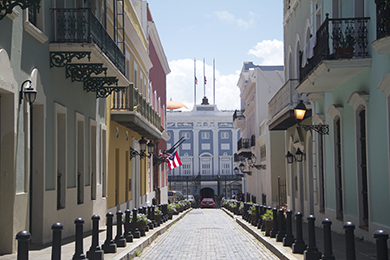 cobbled street lined with colorful colonial buildings in old san juan, puerto rico. the puerto rican flag hangs on the left, with lanterns attached to the buildings. a blue building with flags is visible at the street's end.