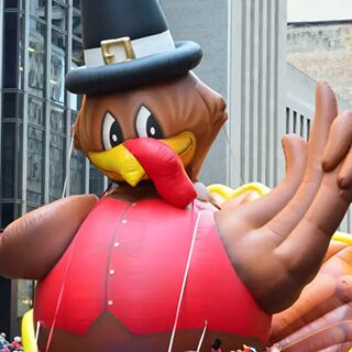 a large inflatable turkey balloon with a black pilgrim hat and red coat, part of a parade. it stands tall against a backdrop of modern buildings. people are gathered below, holding ropes attached to the balloon. a large inflatable turkey balloon with a black pilgrim hat and red coat, part of a parade. it stands tall against a backdrop of modern buildings. people are gathered below, holding ropes attached to the balloon.