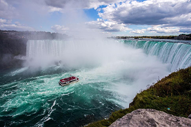 a boat sails near the misty base of a large waterfall, surrounded by lush greenery and under a partly cloudy sky.