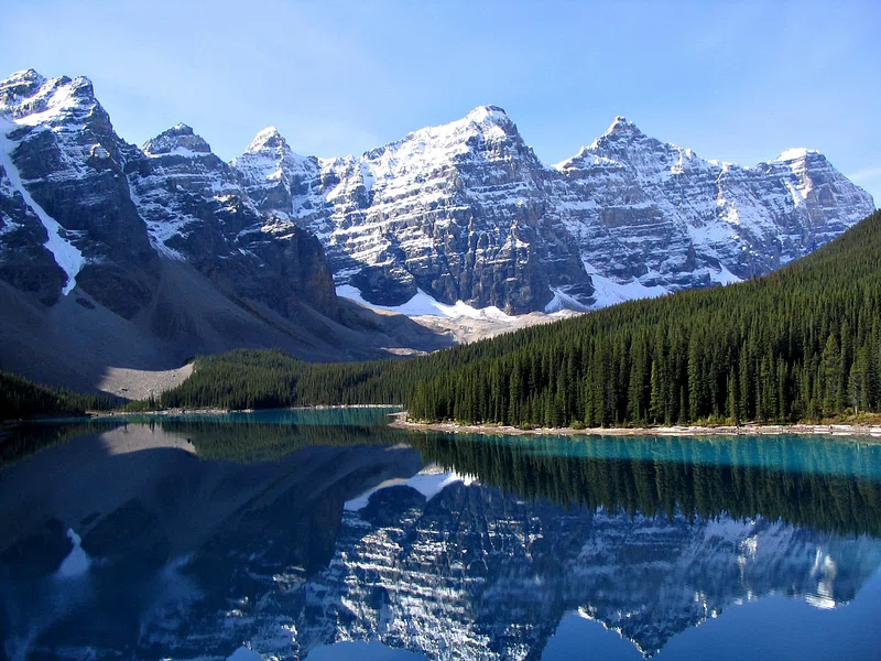 a serene lake reflects snow-capped mountains and a dense pine forest under a clear blue sky. the rugged peaks form a majestic backdrop, creating a mirror-like symmetry on the water's surface.