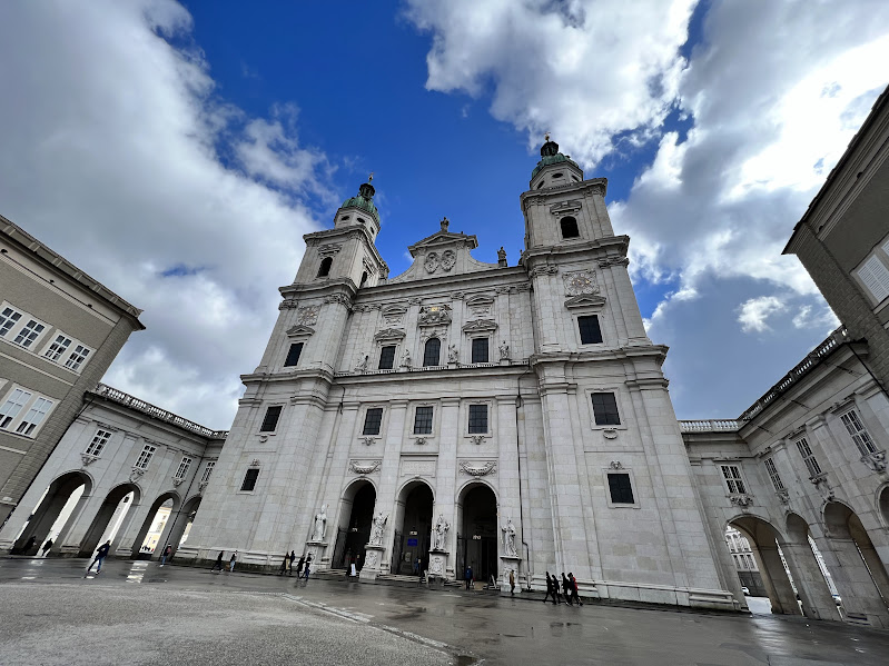 the image shows a large, ornate building with two towers against a partly cloudy sky. the structure is likely a cathedral, featuring arched doorways and intricate architectural details. a few people are walking in the foreground.