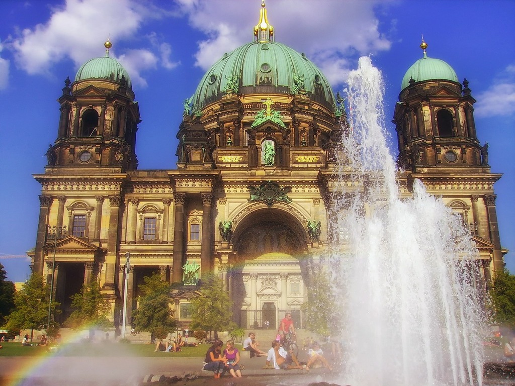 the image shows the berlin cathedral with its ornate baroque architecture and distinctive green domes under a clear blue sky. a fountain in the foreground sprays water, creating a rainbow effect. people are sitting on the grass, enjoying the view.